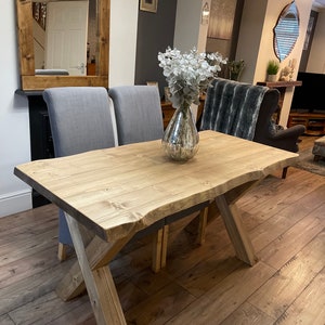 May include: A light-colored wooden dining table with an X-shaped base. The table has a natural wood grain finish and is paired with two gray upholstered chairs. A decorative vase with white flowers sits on the table. The table is approximately 72 inches long.