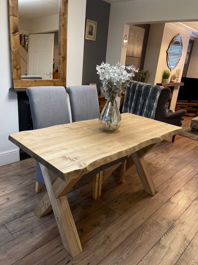 May include: A light-colored wooden dining table with an X-shaped base. The table has a natural wood grain finish and is paired with two gray upholstered chairs. A decorative vase with white flowers sits on the table. The table is approximately 72 inches long.