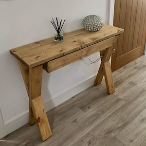 May include: A rustic wooden console table with a single drawer and a criss-cross base. The table is made of natural wood with a light brown finish.