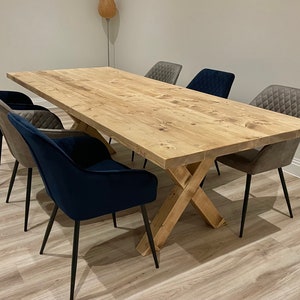 May include: A large wooden dining table with a natural finish and a cross-shaped base. The table is surrounded by six chairs, two with gray fabric upholstery and four with dark blue velvet upholstery.
