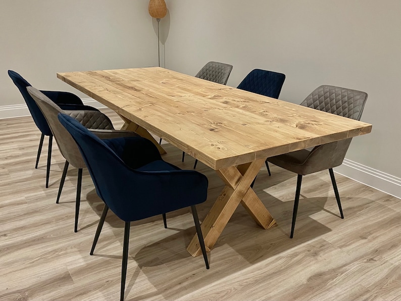 May include: A large wooden dining table with a natural finish and a cross-shaped base. The table is surrounded by six chairs, two with gray fabric upholstery and four with dark blue velvet upholstery.