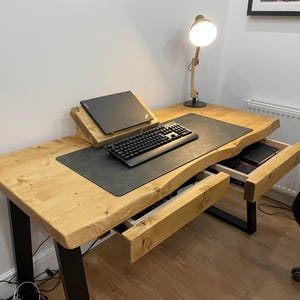 May include: A wooden desk with a black metal frame and two drawers. The desk has a black mousepad and a keyboard on it. A lamp is on the desk.