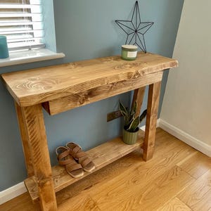 May include: A wooden console table with a natural finish. The table has a live edge top and a lower shelf. A pair of brown sandals rests on the shelf. A small potted plant and a candle are on the table.