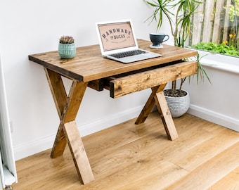 May include: A rustic wooden desk with an X-shaped leg design. The desk has a natural wood finish and a pull-out drawer. A laptop with the text "HANDMADE IN BUCKS" is on the desk, along with a small succulent in a teal pot and a blue mug.