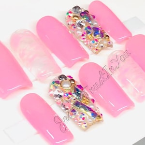 May include: Pink press-on nails with a single accent nail featuring a cluster of iridescent rhinestones, pearls, and gold accents. The text "Jess Trust In You" is visible in the image.