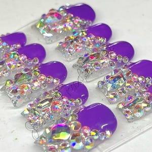 Puede incluir: Un conjunto de uñas postizas con una base morada vibrante y puntas transparentes. Las puntas están profusamente adornadas con una variedad de pedrería iridiscente, creando un efecto brillante. Las uñas están dispuestas sobre una superficie transparente.