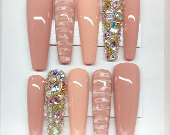 May include: Set of ten long, nude-colored fake nails. Some nails have a white marble design, while others are adorned with rhinestones and pearls.