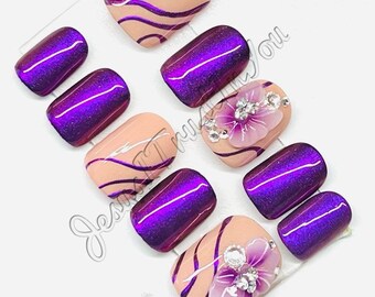 Op de afbeelding: Set van tien kunstnagels met een nude basis en paarse glitter tips. Sommige nagels hebben een roze bloem met strass.