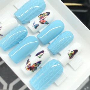 May include: Set of ten blue and white press-on nails with butterfly designs. The nails have a textured surface and are designed to look like a knitted sweater.