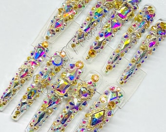 May include: Set of ten clear press-on nails decorated with iridescent rhinestones and gold accents. The nails are arranged in a row on a white background.