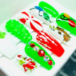 May include: Set of ten Christmas-themed false nails. The nails are decorated with various designs, including candy canes, Christmas lights, a Grinch, and a reindeer. The nails are all green, red, and white.