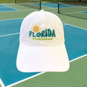 Pode incluir: Bon&eacute; de beisebol branco com um design bordado que apresenta um sol, uma bola de pickleball e o texto "Florida Pickleball".