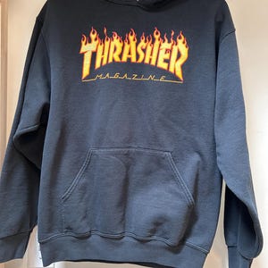 Może przedstawiać: Czarna bluza z kapturem z kieszenią typu kangur. Z przodu znajduje się napis "Thrasher Magazine" w żółtych i pomarańczowych płomieniach. Bluza wisi na wieszaku.