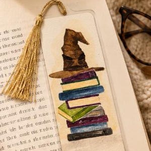 Puede incluir: Un marcapáginas rectangular de papel con una ilustración en acuarela de un sombrero puntiagudo marrón sobre una pila vertical de libros coloridos. El marcapáginas está decorado con una borla dorada metálica.