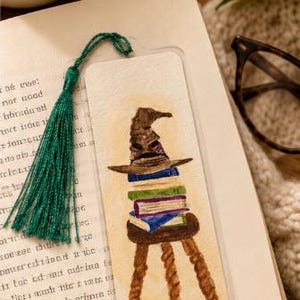 Puede incluir: Un marcapáginas rectangular de papel con una ilustración en acuarela de un sombrero puntiagudo marrón sobre una pila de libros coloridos en un taburete de madera de tres patas. El marcapáginas tiene una borla verde en la esquina.