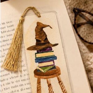 Puede incluir: Un marcapáginas rectangular con una ilustración en acuarela de un sombrero puntiagudo marrón sobre una pila de libros coloridos encima de un taburete de madera. El marcapáginas incluye una borla dorada unida al borde superior.