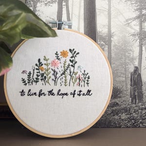 May include: Embroidered floral hoop art featuring a variety of colorful flowers and greenery. The text "to live for the hope of it all" is stitched below the flowers. The embroidery is set in a wooden hoop.