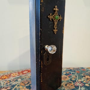 May include: A vintage black wooden box with a decorative brass cross and a green gem in the center. The box has a clear glass knob and a keyhole design. The side of the box is painted silver.