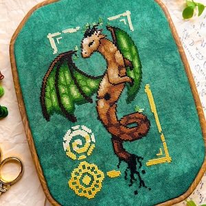 May include: Cross-stitch art of a dragon with green wings and a brown body, set against a teal background. The artwork is framed in a wooden hoop. Decorative scissors and embroidery floss are visible.