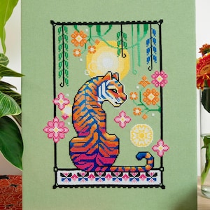 May include: Cross-stitch art of a tiger with blue, orange, and pink stripes, set against a green background. The design includes floral elements and a yellow sun. The artwork is framed in a black border.