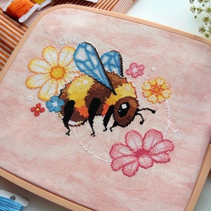 May include: A pink and white embroidery hoop featuring a cross-stitch design of a bee surrounded by flowers. The bee is yellow and black with blue wings. The flowers are pink, white, and yellow.