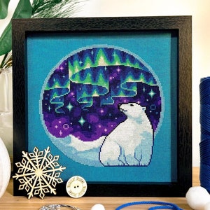 Op de afbeelding: Een ingelijst kruissteekontwerp met een ijsbeer die voor een blauwe en paarse aurora borealis staat. Het ontwerp is omgeven door een witte cirkel en een blauwe achtergrond.