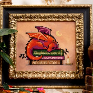 May include: Cross-stitch art of a red and purple dragon resting on books titled "Flight Manual", "Alchemy 101", and "Ancient Tales". The framed artwork has a gold ornate frame and a black inner frame. A small candle is lit.