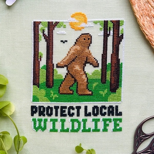 Puede incluir: Arte de punto de cruz que representa un Bigfoot marrón caminando en una escena de bosque con un sol amarillo. El texto "PROTECT LOCAL WILDLIFE" está bordado en verde y blanco. La imagen está rodeada de hilo verde, naranja, marrón y unas tijeras.