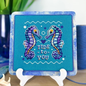 May include: Cross-stitch art featuring two seahorses facing each other, with the words "Tide to You" stitched below. The seahorses are purple and blue on a turquoise background, framed in a blue marbled border. The artwork is displayed on a white easel.