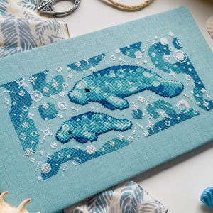 May include: A rectangular light blue fabric panel featuring a cross-stitch embroidery of two manatees surrounded by white and dark blue geometric shapes, stars, and bubbles. The design is centred on the fabric, showcasing detailed needlework.