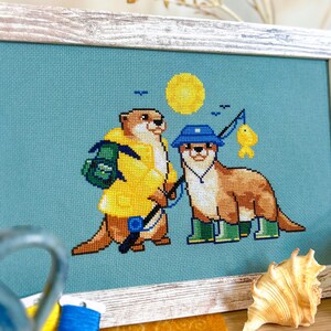May include: Cross-stitch artwork in a wooden frame. The image shows two otters: one in a yellow raincoat and backpack, the other fishing with a blue hat and wellington boots. A yellow sun and blue background complete the scene.