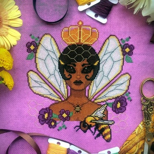 Vertical front view of Queen Bee cross stitch pattern. Queen Bee is represented as a black woman with dark hair that is cut into a bob. She is wearing an orange crown and has the wings of a bee. She wears a necklace and is surrounded by flowers.