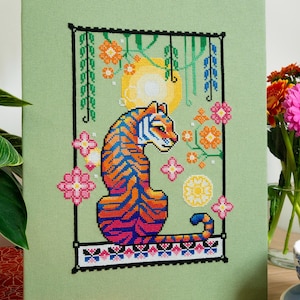 May include: Cross-stitch art of a tiger with orange and blue stripes, set against a light green background. The design includes flowers, a sun, and a decorative border. The artwork is framed and displayed on a wooden surface.