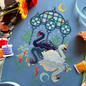 May include: Cross-stitch embroidery of two swans, one black and one white, under a stained glass archway with a crescent moon. The embroidery is surrounded by green foliage and red flowers.