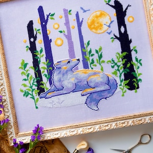 Peut inclure: Une œuvre d'art au point de croix dans un cadre doré représente un loup dans une scène de forêt. Le loup est bleu et jaune, couché devant des arbres, une pleine lune et des oiseaux. L'œuvre est sur tissu blanc. Le cadre est orné.