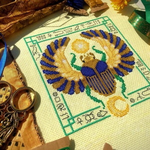 Peut inclure: Une broderie au point de croix représentant un scarabée ailé, entouré de hiéroglyphes. Le scarabée est bleu et or, avec un soleil jaune derrière lui. La broderie est sur un tissu blanc avec une bordure verte.