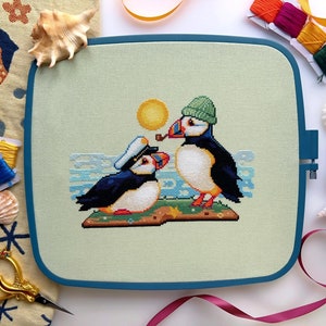May include: Cross-stitch embroidery of two puffins on a green background. The puffin on the left is wearing a blue sailor hat and the puffin on the right is wearing a green hat. The puffin on the right is holding a pipe in its beak.