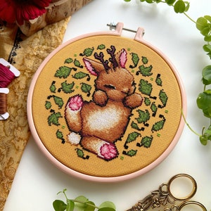 May include: A cross-stitch embroidery hoop with a light brown deer with antlers curled up in a circle of green leaves on a yellow background.