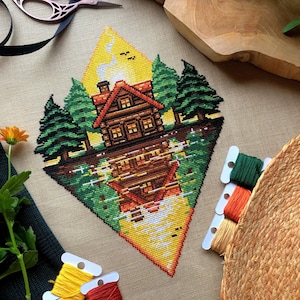 Może przedstawiać: Haftowane krzyżykiem przedstawienie chatki w lesie, z odbiciem chatki w wodzie poniżej. Chatka jest brązowa z czerwonym dachem i kominem, z którego wydobywa się dym. Drzewa są zielone, a woda niebieska. Tło stanowi żółty romb.