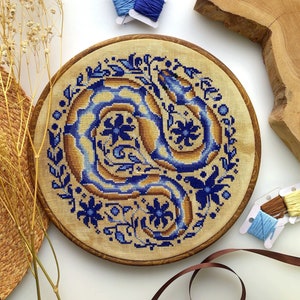 Peut inclure: Un cercle de broderie circulaire avec un motif floral bleu et or repr&eacute;sentant un serpent stylis&eacute;. Le motif est sur un fond de tissu beige.