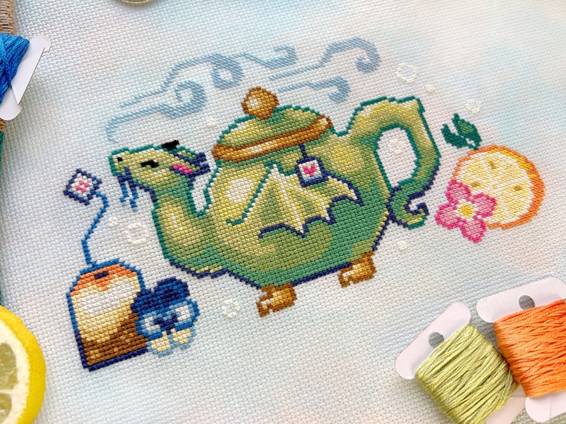 May include: A green teapot with a dragon handle and a tea bag hanging from the spout. The teapot is surrounded by a lemon, a flower, and a tea bag. The background is a light blue and white watercolor wash.