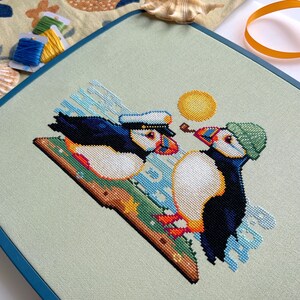 May include: Cross-stitch pattern of two puffins on a green and blue background. One puffin is wearing a blue and white sailor hat and the other is wearing a green hat. The sun is shining in the background.