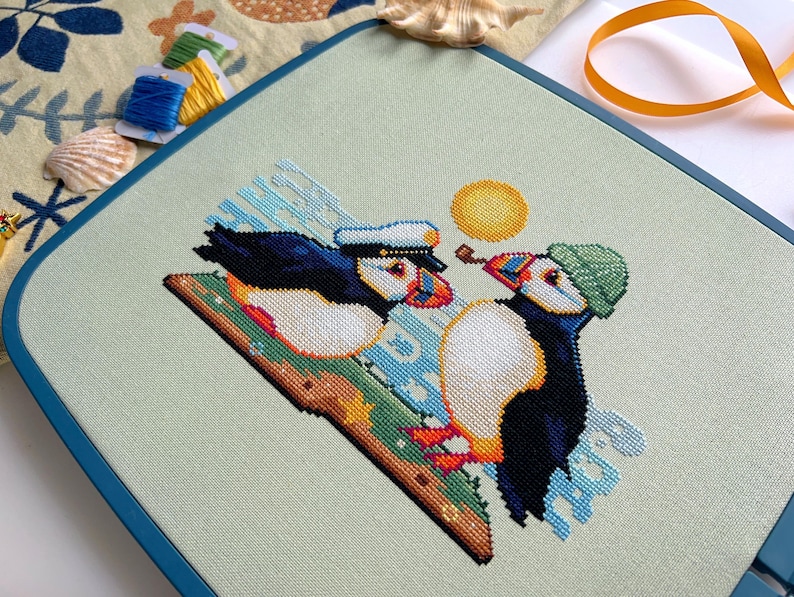 May include: Cross-stitch pattern of two puffins on a green and blue background. One puffin is wearing a blue and white sailor hat and the other is wearing a green hat. The sun is shining in the background.