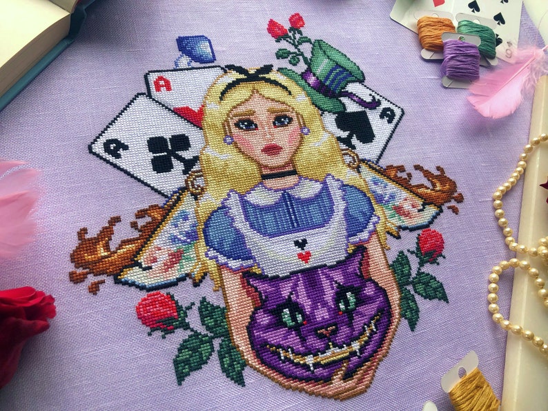 Front, angled view of Alice and Cheshire Cat cross stitch pattern. Cheshire cat head is purple and large, with bared teeth and green eyes. Alice is surrounded by objects like cards, a hat, teacups, roses and a mushroom.