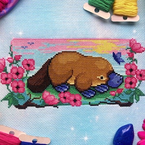Closeup of Platypus cross stitch pattern, scene surrounded by flowers and bobbins. Colors are bright. Mainly pink, blue, yellow, green, and brown are present. Platypus is facing right, and is surrounded by pink flowers. There is a blue butterfly.