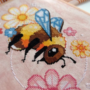 May include: A cross-stitch embroidery of a bee with yellow and brown stripes, blue wings, and black legs. The bee is surrounded by flowers in shades of pink, yellow, and blue.