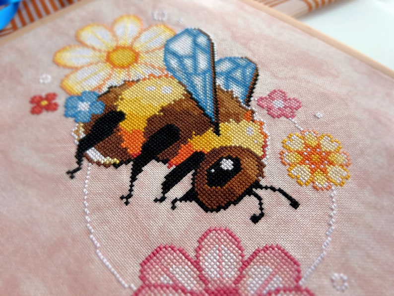 May include: A cross-stitch embroidery of a bee with yellow and brown stripes, blue wings, and black legs. The bee is surrounded by flowers in shades of pink, yellow, and blue.