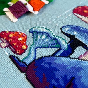 May include: Close-up of a cross-stitch embroidery project depicting a whimsical scene with mushrooms in shades of blue, green, and red. The embroidery is on a light blue fabric.