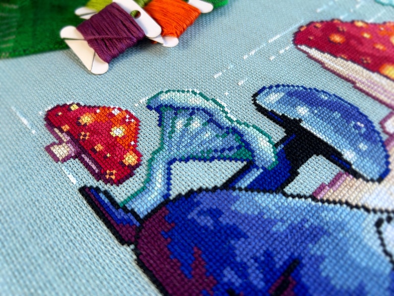 May include: Close-up of a cross-stitch embroidery project depicting a whimsical scene with mushrooms in shades of blue, green, and red. The embroidery is on a light blue fabric.