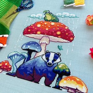 May include: A cross-stitch embroidery featuring a badger standing under a red and yellow mushroom with a green frog on top. The mushroom is surrounded by other mushrooms in shades of blue, green, and yellow. The background is a light blue canvas with white raindrops.
